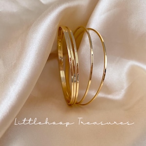 Puede incluir: Un conjunto de tres brazaletes finos de metal en tono dorado pulido con una superficie lisa y reflectante. Las pulseras están dispuestas en grupo, mostrando su forma circular y brillo metálico. El texto Littlehoop Treasures es visible en la parte inferior.