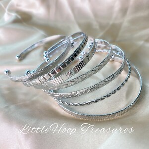 May include: Set of six silver bangles with different engraved patterns. The bangles are stacked on top of each other, showcasing the intricate designs. The text "Little Hoop Treasures" is visible at the bottom of the image.