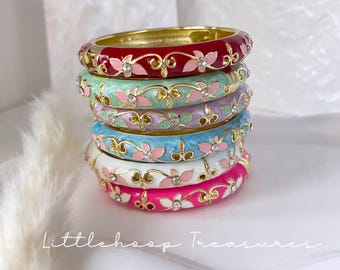Op de afbeelding: Een stapel van zes scharnierende armbanden in rood, mintgroen, lavendel, lichtblauw, wit en felroze. Elke metalen armband is voorzien van goudkleurig filigraan, roze emaille bloemblaadjes en kleine, ingebedde doorzichtige strass-steentjes.
