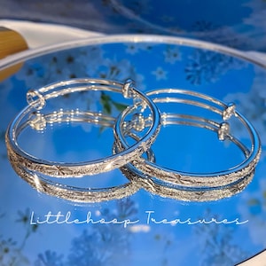 Könnte beinhalten: Zwei silberfarbene, verstellbare Armreifen mit einer texturierten, glitzernden Oberfläche und eingravierten sternartigen Mustern. Die Armreifen sind kreisförmig und haben überlappende Enden. Der Text Littlehoop Treasures ist unten zu sehen.