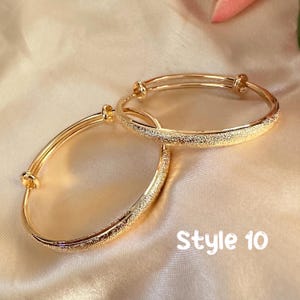 May include: Two gold-colored adjustable bangles with a textured, glittery band. The bangles are displayed on a satin-like surface. The text "Style 10" is visible in the lower right corner. These bangles are a type of jewelry.