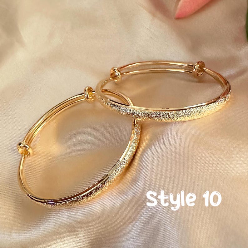 May include: Two gold-colored adjustable bangles with a textured, glittery band. The bangles are displayed on a satin-like surface. The text "Style 10" is visible in the lower right corner. These bangles are a type of jewelry.