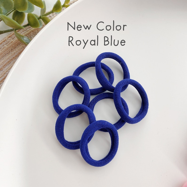 May include: A close-up of a pile of royal blue hair elastics on a white surface. The text "New Color Royal Blue" is written above the elastics.