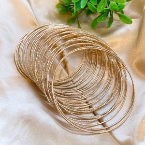 May include: A set of multiple thin, circular gold-toned bangles. Each bangle features a delicate, textured surface with small, repeating indentations that catch the light. The bracelets are stacked together, creating a layered, metallic appearance.