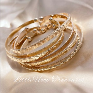 May include: A set of six gold-toned bangle bracelets with different textured patterns. The bracelets are stacked on top of each other and have a shiny finish.