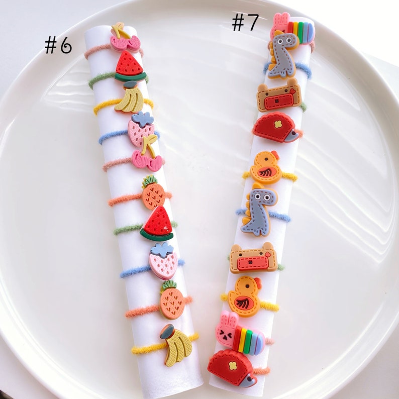 May include: Two sets of colorful hair ties with various cartoon charms. The set labeled #6 features fruit charms like watermelon, pineapple, and bananas. The set labeled #7 features animal charms like a dinosaur, a chick, and a hedgehog.