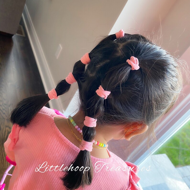May include: A young girl with long dark hair styled with multiple pink hair ties. The hair is pulled back in a series of ponytails. The girl is wearing a pink shirt and a colorful beaded necklace. The text "Littlehoop Treasures" is visible in the bottom right corner of the image.