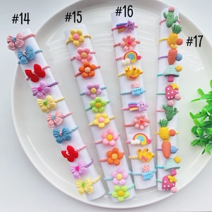 May include: A set of 17 colorful hair ties with various cartoon designs, including flowers, rainbows, stars, and a pineapple. The hair ties are arranged in rows on a white plate, labeled with numbers 14 through 17.