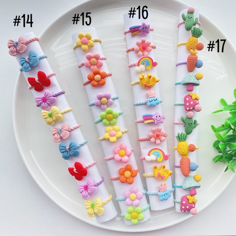 May include: A set of 17 colorful hair ties with various cartoon designs, including flowers, rainbows, stars, and a pineapple. The hair ties are arranged in rows on a white plate, labeled with numbers 14 through 17.