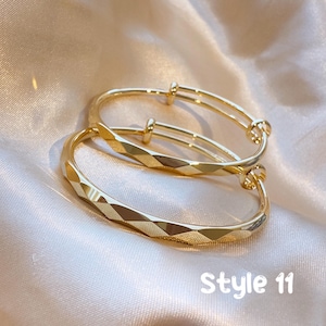 May include: Two gold-colored adjustable bracelets. The bracelets have a geometric diamond pattern on the front and a smooth, rounded design on the sides. The text "Style 11" is visible in white.