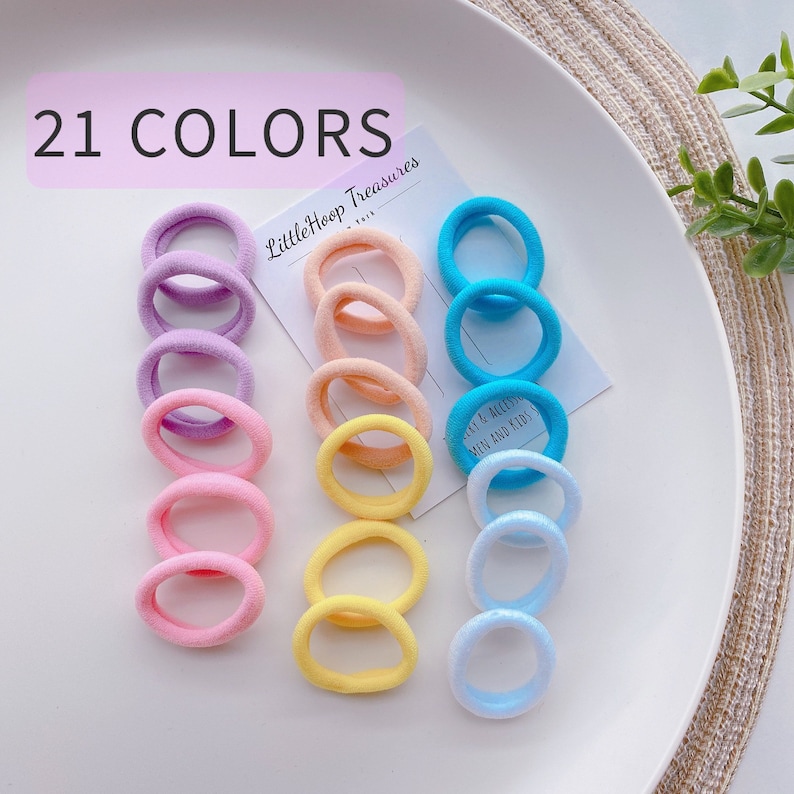 May include: A set of 21 colorful hair ties in various shades of pink, purple, blue, yellow, and orange. The hair ties are arranged in groups of three on a white plate. The text "21 COLORS" and "Littlehoop Treasures" is visible in the image.