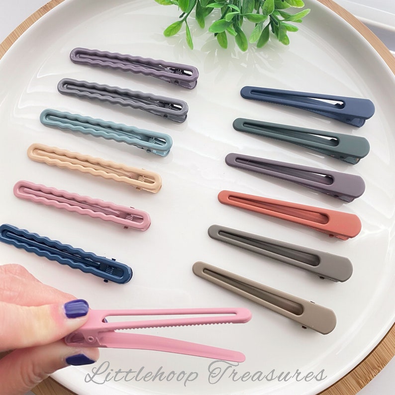 Op de afbeelding: Assortiment van 12 kleurrijke plastic haarclips. De clips zijn in verschillende tinten blauw, paars, roze, bruin en grijs. De clips zijn allemaal even groot en hebben dezelfde vorm, met een gladde, matte afwerking.