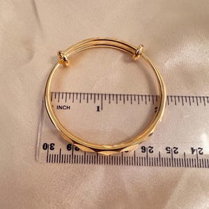 May include: A gold-colored adjustable bangle bracelet with a geometric design. The bracelet is measured with a ruler in inches. The bracelet is on a light beige satin surface.