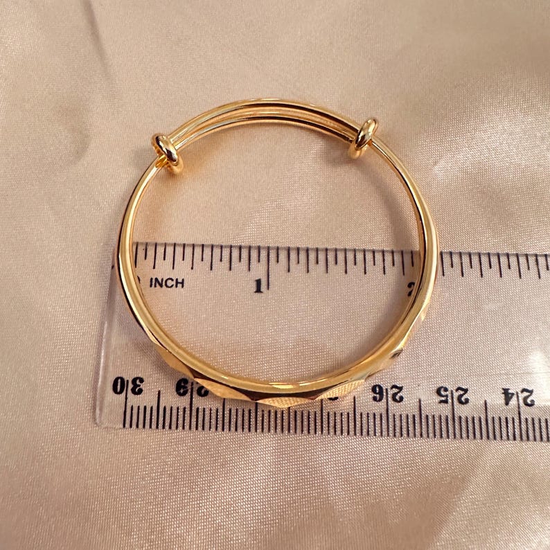 May include: A gold-colored adjustable bangle bracelet with a geometric design. The bracelet is measured with a ruler in inches. The bracelet is on a light beige satin surface.