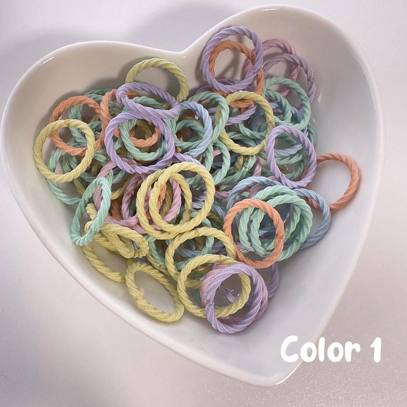 May include: A white heart-shaped bowl filled with a variety of pastel-colored rope rings. The rings are in shades of light blue, light green, light pink, and yellow. The text "Color 1" is written in black at the bottom of the image.