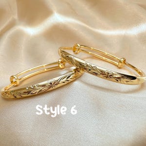 May include: Two gold-colored adjustable bangles with floral and leaf patterns. The bangles are displayed on a satin surface. The text "Style 6" is visible in the image.