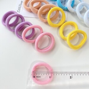May include: A set of colorful hair ties in various pastel colors, including purple, pink, yellow, and blue. The hair ties are arranged on a white surface with a ruler showing the size of one of the hair ties is 1 inch.