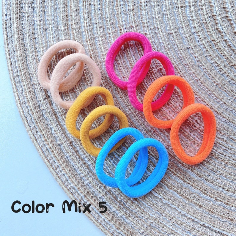 May include: A set of 10 colorful hair ties in a rainbow of colors, including pink, orange, yellow, blue, and peach. The hair ties are arranged in a circle on a woven surface. The text "Color Mix 5" is visible in the image.