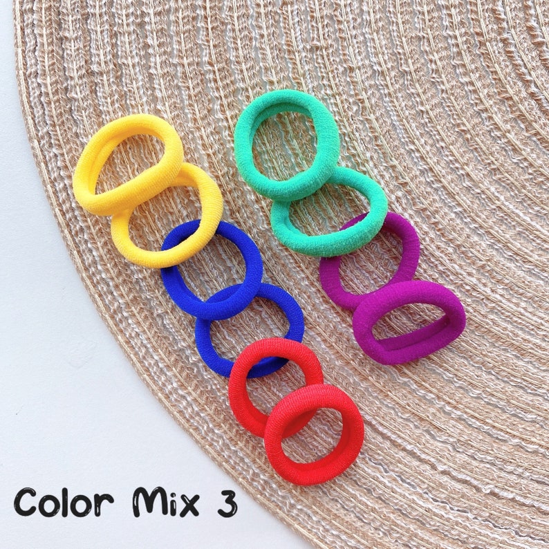 May include: A set of 8 colorful hair ties in yellow, green, blue, and red. The hair ties are arranged on a woven mat.
