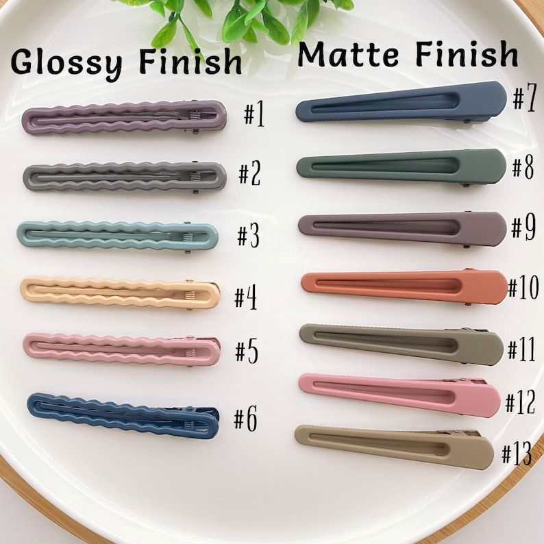 Op de afbeelding: Twee rijen haarclips, &eacute;&eacute;n rij met een glanzende afwerking en &eacute;&eacute;n rij met een matte afwerking. De clips met een glanzende afwerking hebben een golvend ontwerp en zijn verkrijgbaar in zes kleuren: paars, grijs, teal, beige, roze en blauw. De clips met een matte afwerking zijn recht en zijn verkrijgbaar in zeven kleuren: blauw, groen, grijs, oranje, groen, roze en bruin.