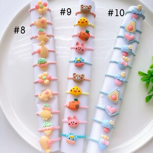 May include: Set of 18 colorful hair ties with cute cartoon animal and food charms. The hair ties are arranged in three rows of six, labeled #8, #9, and #10. Each hair tie features a different charm, including a pineapple, a flower, a cupcake, and a heart.