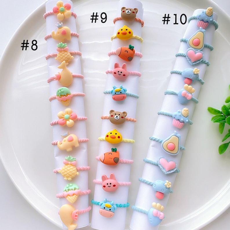 May include: Set of 18 colorful hair ties with cute cartoon animal and food charms. The hair ties are arranged in three rows of six, labeled #8, #9, and #10. Each hair tie features a different charm, including a pineapple, a flower, a cupcake, and a heart.