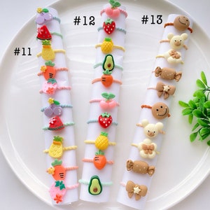 May include: A set of 12 colorful hair ties with various fruit and animal designs. The hair ties are arranged in three rows of four, with each row labeled with a number. The hair ties are made of elastic and have a small charm attached to each one. #11, #12, #13