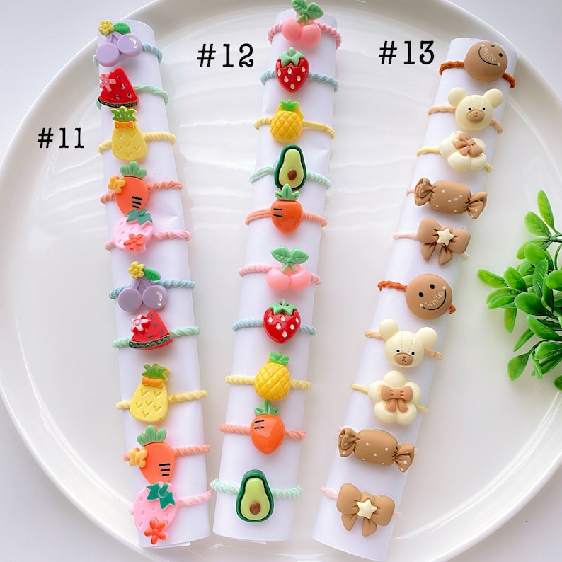 May include: A set of 12 colorful hair ties with various fruit and animal designs. The hair ties are arranged in three rows of four, with each row labeled with a number. The hair ties are made of elastic and have a small charm attached to each one. #11, #12, #13