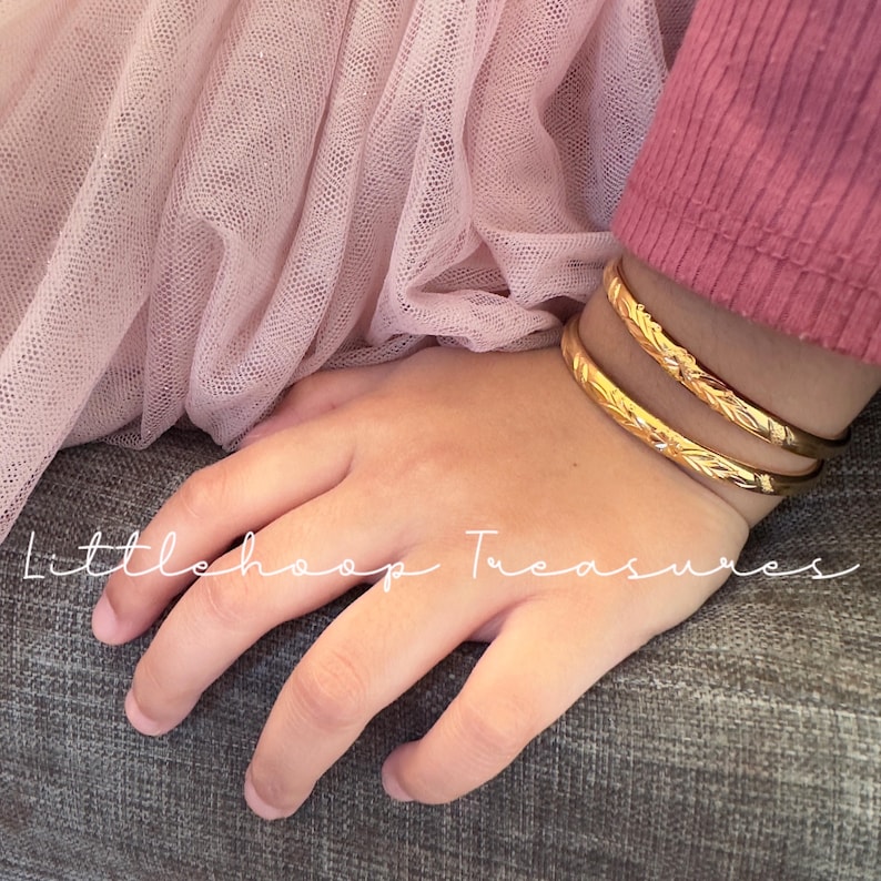 May include: Two gold-colored bangles with leaf-like patterns are worn on a hand. The hand is resting on a gray surface, with a pink tulle fabric and a pink long-sleeved shirt in the background. The text "Littlehoop Treasures" is written on the image.
