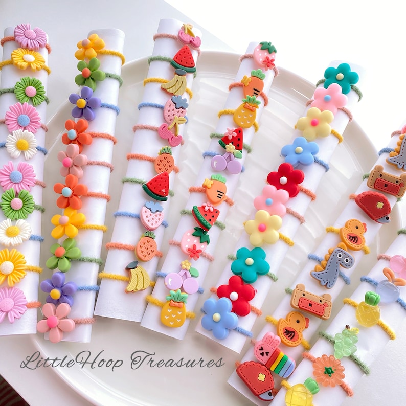 May include: A set of colorful hair ties with various cute charms, including flowers, fruits, and animals. The hair ties are made of elastic and are perfect for holding back hair.