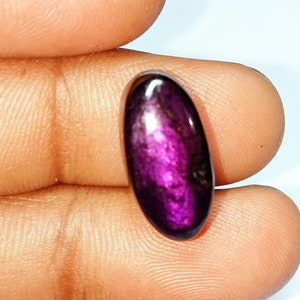 Pode incluir: Uma gema oval de cor p&uacute;rpura profunda com um efeito brilhante e iridescente. A pedra &eacute; segurada na m&atilde;o, mostrando seu tamanho e cor.
