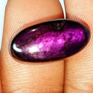 Pode incluir: Uma gema oval caboch&atilde;o de cor roxa e preta com um acabamento brilhante e iridescente.