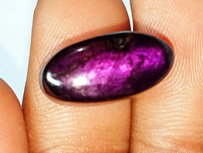 Pode incluir: Uma gema oval caboch&atilde;o de cor roxa e preta com um acabamento brilhante e iridescente.