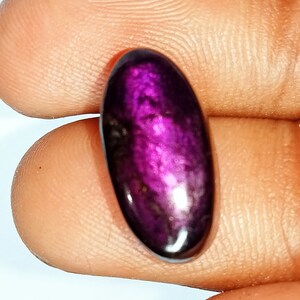 Pode incluir: Um close-up de uma gema oval de cor p&uacute;rpura com um acabamento brilhante e iridescente. A gema &eacute; segurada na m&atilde;o, mostrando sua cor e textura &uacute;nicas.