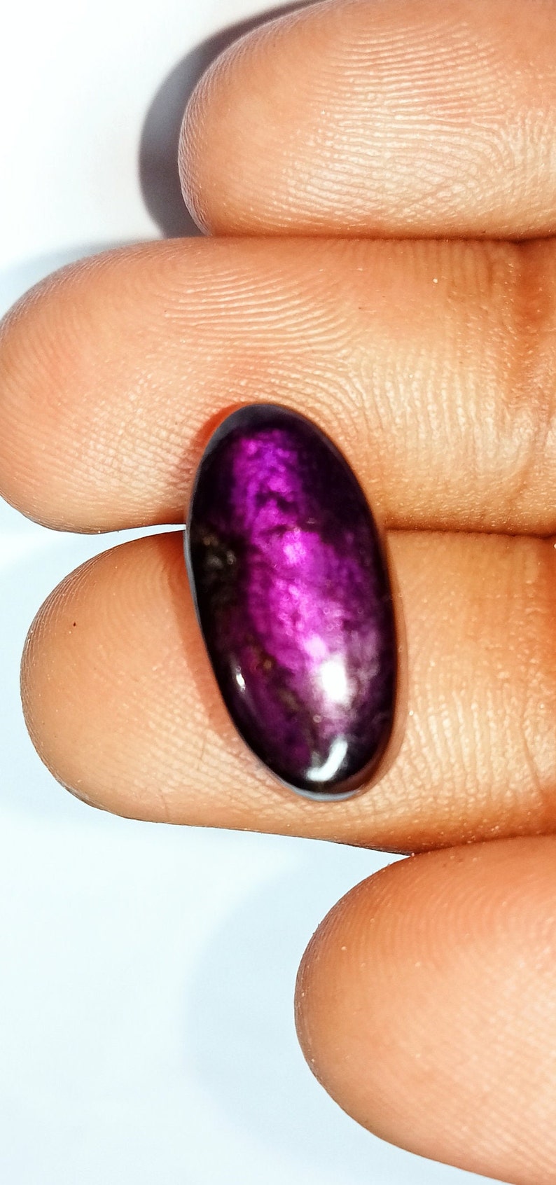 Pode incluir: Um close-up de uma gema oval de cor p&uacute;rpura com um acabamento brilhante e iridescente. A gema &eacute; segurada na m&atilde;o, mostrando sua cor e textura &uacute;nicas.