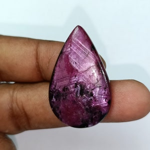 Può includere: Una pietra preziosa rosa scuro a forma di lacrima con un motivo a stella. La pietra preziosa ha un bordo scuro, quasi nero, attorno ai bordi.