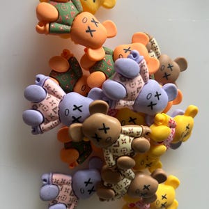May include: A collection of small, flat bear-shaped charms in various colours including orange, yellow, brown, and lavender. Each bear features a printed pattern of small symbols and has "X" marks for eyes and a simple line for a mouth on its face.