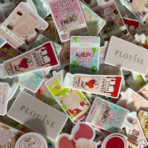 May include: A collection of assorted stickers featuring Plouise cosmetic products, including rectangular logos, tubes of paint, and drink-shaped designs with text like Bad Bitch Energy and Juicy Drip in various pastel and vibrant colours.