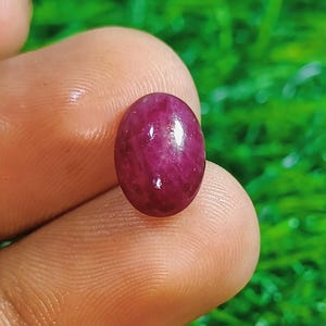 May include: A polished, oval-shaped ruby gemstone, held between fingers, showcasing a deep red hue with subtle internal patterns. The gem is approximately 0.5 inches in length, set against a blurred green background.