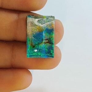 May include: A rectangular opal gemstone with a vibrant display of color. The stone features a mix of blue, green, and gold hues, with a translucent appearance. The opal is held between fingers against a white background.