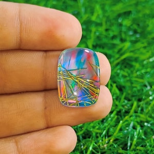 Könnte beinhalten: Ein rechteckiger Edelstein mit transluzenter Basis, der interne Strukturen aus Gold und Grün mit roten und blauen Streifen aufweist. Der Stein wird in einer Hand vor grünem Gras gehalten.