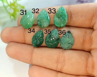 Puede incluir: Seis colgantes de piedras preciosas verdes talladas, numerados del 31 al 36, se muestran en una mano. Los colgantes tienen forma de hoja con intrincados detalles tallados. El fondo incluye follaje verde y flores moradas.