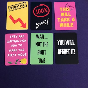 May include: A collection of six colorful cards with text. The cards have phrases like "NEGATIVE," "100% yes!," and "YOU WILL REGRET IT!" The cards are arranged on a dark purple surface.