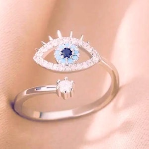 May include: Silver adjustable ring with an eye design. The eye features a blue and white colour scheme with a dark blue centre, surrounded by clear crystals. The ring's band is a simple, polished metal.