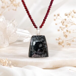 May include: A necklace featuring a trapezoidal pendant with a dark grey and black mottled stone set in a silver-toned frame, suspended from a strand of small, faceted, deep red beads that catch the light with a polished finish.