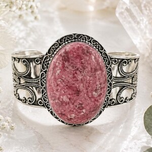 May include: A silver cuff bracelet with an oval, pink gemstone. The gemstone is set in a decorative frame, and the cuff has ornate scrollwork. The bracelet is displayed on a white surface with floral accents.