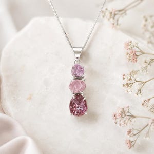 May include: A silver pendant necklace with three pink gemstones of different shades. The top stone is light pink, the middle is medium pink, and the bottom is a deep reddish-pink. The gemstones are set in silver and suspended from a fine silver chain.