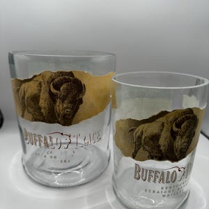 May include: Two clear glass tumblers with a Buffalo Trace design. Each glass features a brown illustration of a bison and the brand name in brown lettering. The glasses are cylindrical and appear to be empty.