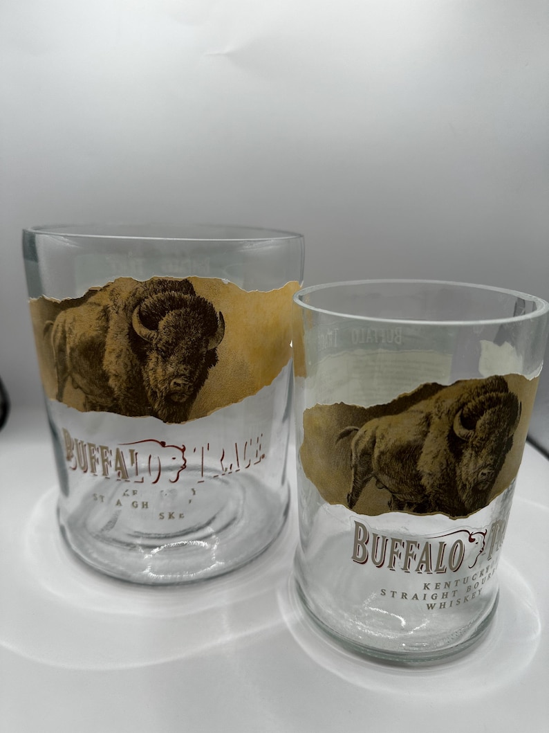 May include: Two clear glass tumblers with a Buffalo Trace design. Each glass features a brown illustration of a bison and the brand name in brown lettering. The glasses are cylindrical and appear to be empty.