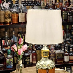 May include: A unique lamp made from a repurposed liquor bottle, filled with small stones, topped with a white lampshade. The bottle has a label that reads "Grand-Dad 86". A small vase with pink tulips sits nearby.